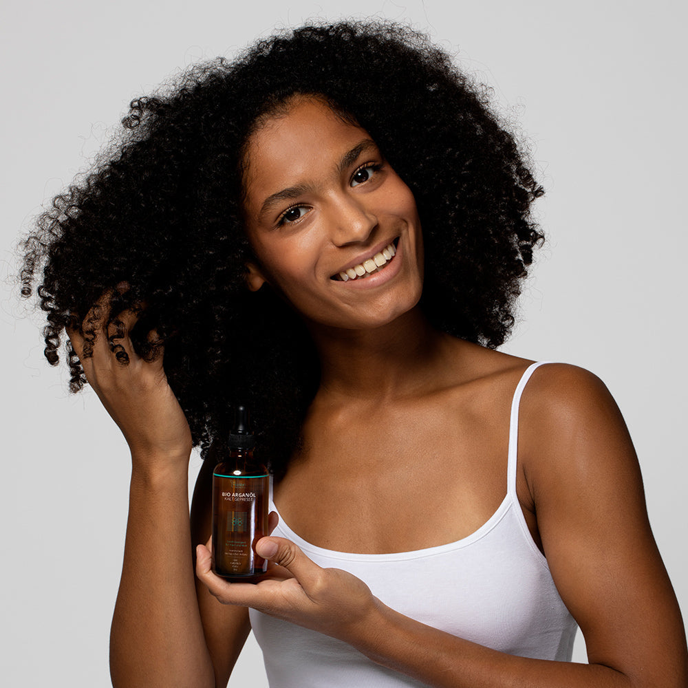 Frau mit lockigem Haar hält eine Flasche Haaröl in der Hand und lächelt in die Kamera.