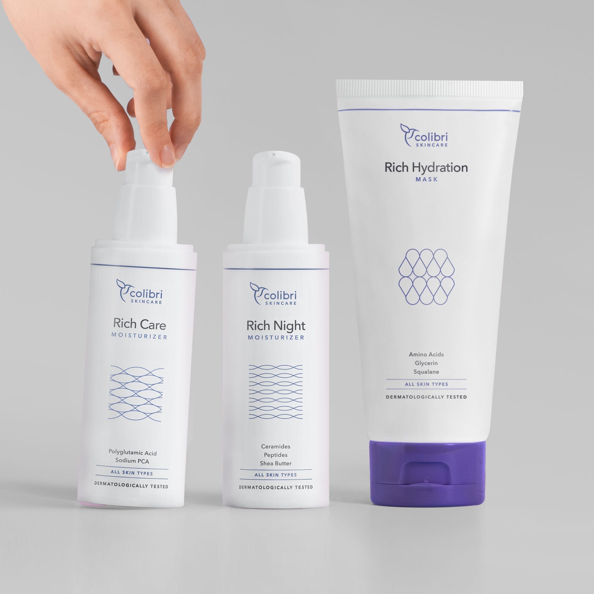 Eine Hand berührt die Pumpflasche eines von drei Hautpflegeprodukten von Colibri Skincare auf grauem Hintergrund.