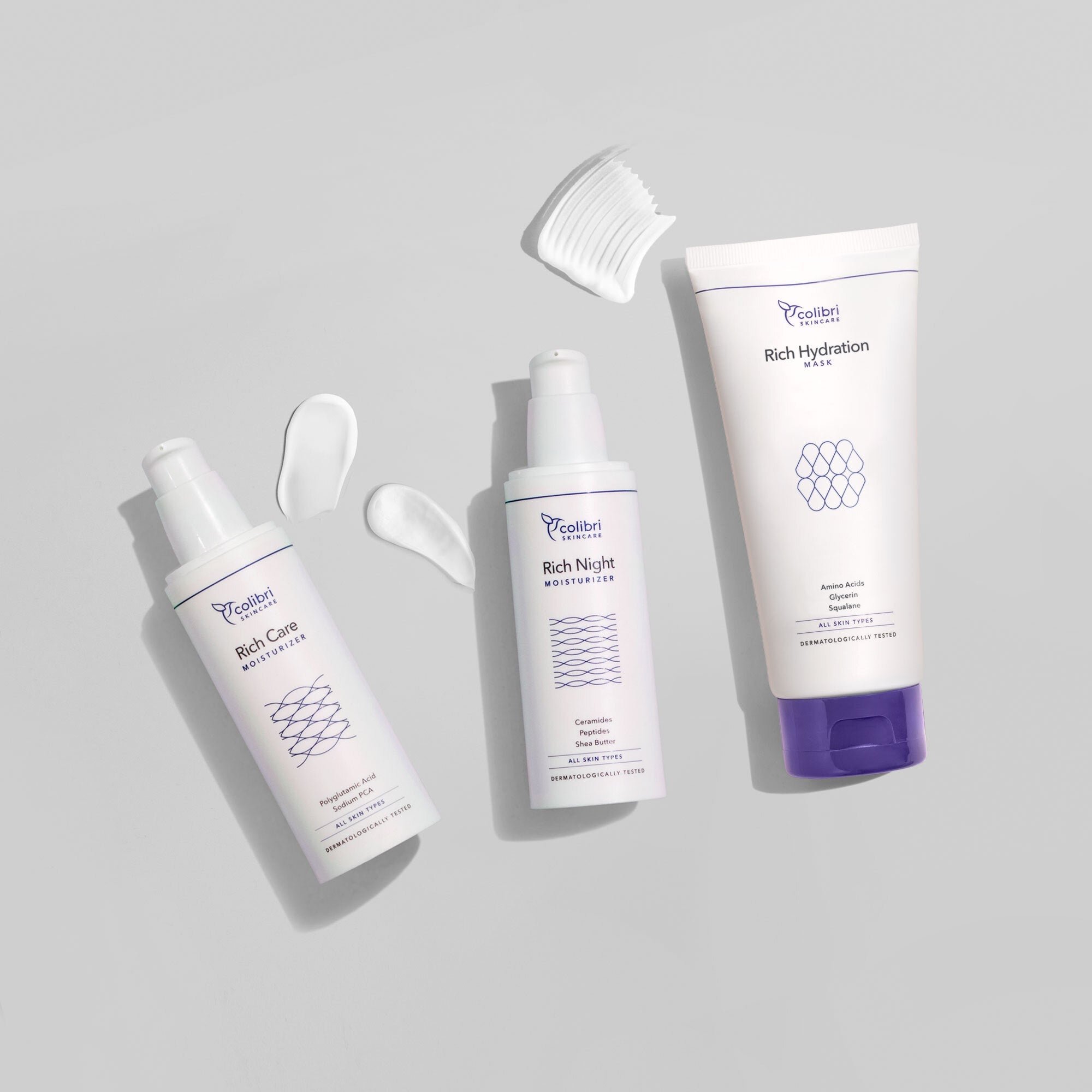 Drei Hautpflegeprodukte auf grauem Hintergrund mit weißen Creme-Tupfern und einer Applikatorbürste.