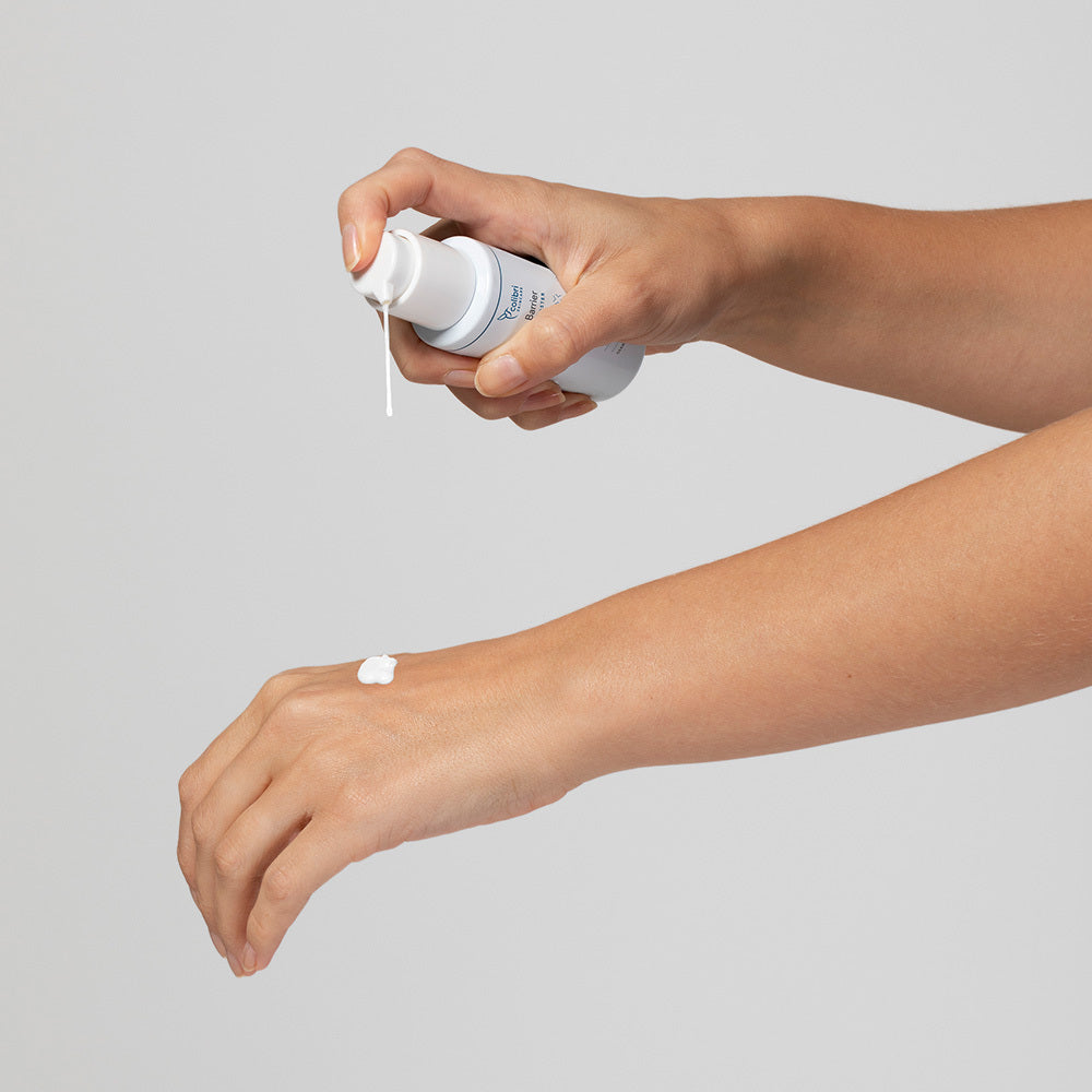 Eine Hand träufelt Lotion aus einer Pumpflasche auf den Handrücken einer anderen Hand vor grauem Hintergrund.