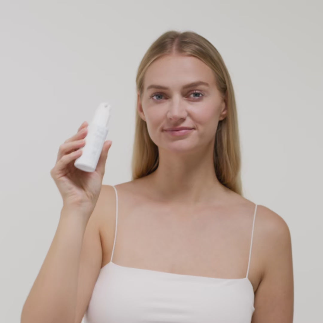Eine Frau in einem weißen Top hält eine Flasche Hautpflegeprodukt vor einem neutralen Hintergrund in der Hand.