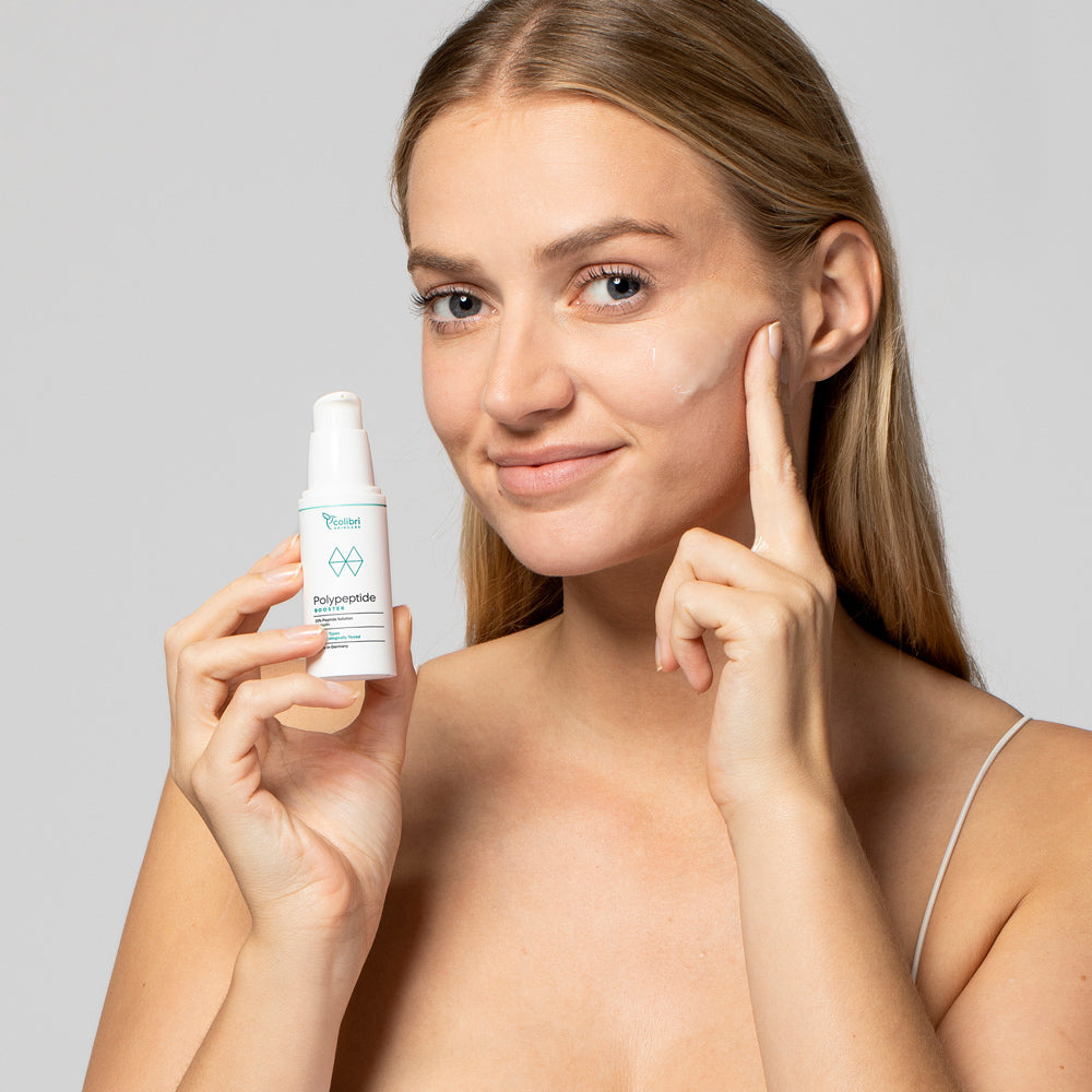 Frau trägt Gesichtsserum auf ihre Wange auf und hält die Flasche in der anderen Hand gegen einen grauen Hintergrund.