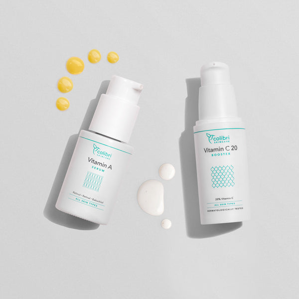 Zwei Fläschchen Hautserum mit Vitamin A und Vitamin C20, umgeben von kleinen Tropfen auf einem grauen Hintergrund.