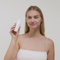 Eine Frau in einem weißen Top hält eine Flasche Hautpflegeprodukt vor einem neutralen Hintergrund in der Hand.